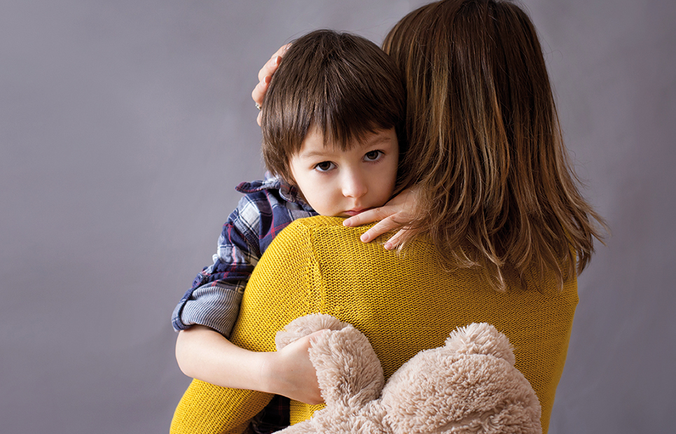 Ein Junge ist auf dem Arm seiner Mutter. Sie steht mit dem Rücken zur Kamera, der Junge umarmt seine Mutter und schaut traurig in die Kamera. In der anderen Hand hält er einen Teddybär fest.