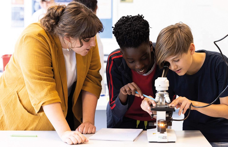 Man sieht zwei Schüler mit der Lehrerin am Mikroskop