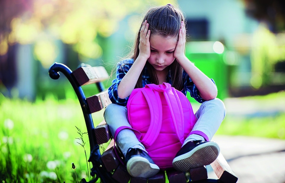 Man sieht ein Schulmädchen, dass mit eihrem Schulranzen auf einer Parkbank sitzt und den Kopf verzweifelt in ihre Hände stützt